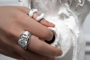 An elegant female hand wearing a scintillating white color Radiant-Cut Moissanite Diamond Ring by www.jingyayi.com.  Well textured artistic organic sculpture in the background.